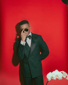 Style influencer Igee Okafor showcases the Paul sunglasses in a stunning formal portrait against a dramatic red backdrop. These refined aviator-style frames in sleek black complement his emerald green double-breasted tuxedo, demonstrating their versatility for formal occasions. The classic design paired with fashion-forward elements creates an iconic look that enhances any formal wear. Handcrafted in Japan with exceptional attention to detail, each pair exemplifies sophisticated style and timeless elegance. The clean lines and luxurious finish make these frames particularly striking for black-tie events and special occasions. Set against white orchids and a rich red background, the sunglasses showcase their ability to enhance formal styling Credits: @igeeokafor