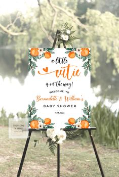 an orange and white wedding sign with greenery on the bottom, in front of a lake