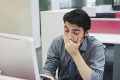 a man sitting at a desk with his hand on his face while looking at a laptop