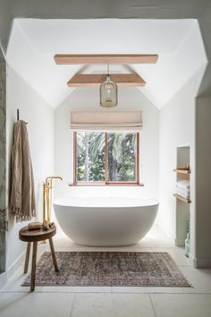 a large white bathtub sitting under a window next to a wooden table and stool