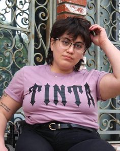 Photo features an young woman wearing a mauve shirt that has our Tinta logo across the front, black jeans, and black belt. She has a mullet, glasses, gold necklace, tattoos on her arm, and is seated in a wheelchair. Mullet Girl, Alternative Girl, Tattooed Girl, Girl Mullet, Alternative Girls, Inspired Fashion, Neck Tattoo, Ethical Fashion, Girl Tattoos