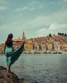 a woman standing on the edge of a body of water with buildings in the background