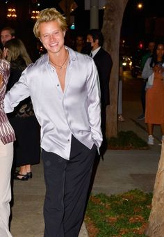 a man in a silver shirt and black pants walking down the street with his hand on his hip