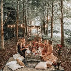 three women sitting at a picnic table in the woods with lights strung over them and talking to each other