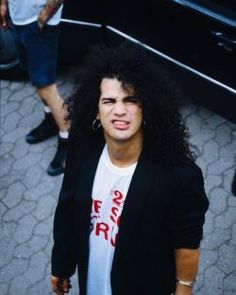 a man with long hair and piercings standing in front of a black car, looking at the camera