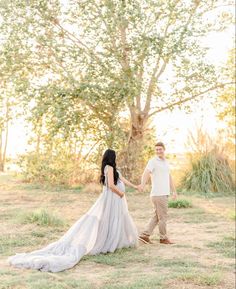 an engaged couple holding hands and walking through the grass in front of a large tree