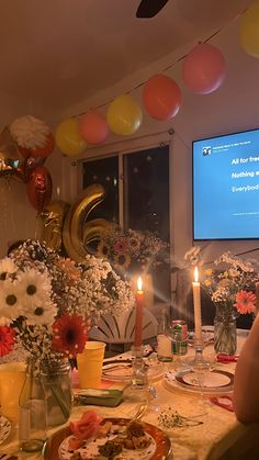 a group of people sitting around a table with food and drinks on it, in front of a flat screen tv