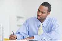 a man sitting at a desk writing on a piece of paper while holding a pen