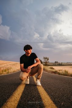 a young man crouches down on the side of an empty road in front of a cloudy sky