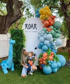 two children are sitting in front of a sign with balloons on it and an animal