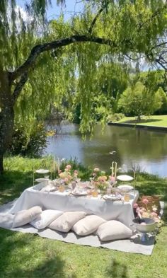 an outdoor table set up with pillows and flowers on the grass next to a lake