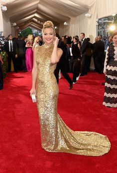 a woman in a gold gown posing on the red carpet at an event with her hand up to her face