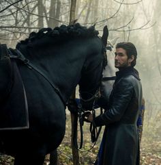 a man standing next to a black horse in the middle of a forest filled with trees