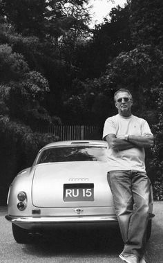 a man leaning on the hood of a sports car in front of trees and bushes