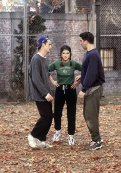 three people standing in front of a chain link fence with leaves on the ground around them