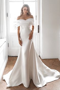 a woman standing in front of a window wearing a wedding dress with off the shoulder sleeves