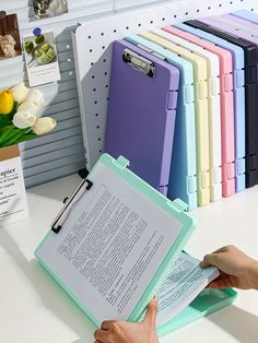 a person is holding a clipboard in front of a book on a table with several different colored folders