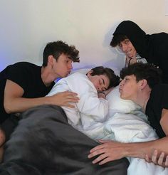 four young men in black hoodies are laying on a bed and one man is covering his face with a blanket