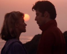 a man and woman standing next to each other near the ocean at sunset or sunrise