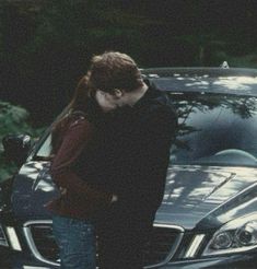 two people standing next to each other in front of a car and one person leaning on the hood