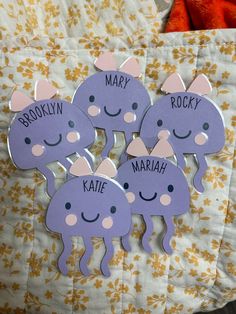 four purple stickers with names on them sitting on a bed next to a stuffed animal