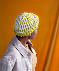 a woman wearing a yellow and white knitted hat with stripes on the front, standing in front of an orange backdrop