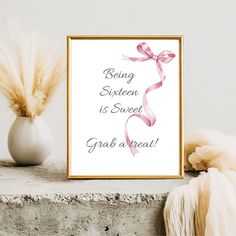 a white vase with flowers and a pink ribbon on it next to a framed print