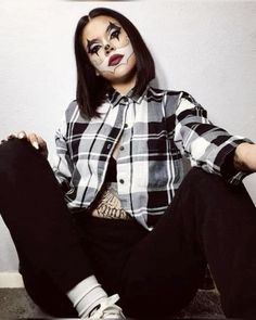 a woman with painted face sitting on the ground wearing black pants and a plaid shirt