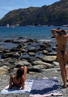 two women in bikinis sitting on rocks near the ocean with boats and mountains behind them