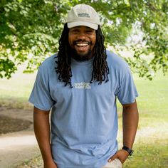 a man with long dreadlocks standing in front of a tree wearing a blue shirt and white hat