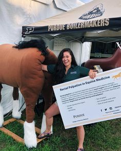 a woman standing next to a brown horse holding a sign in front of a tent