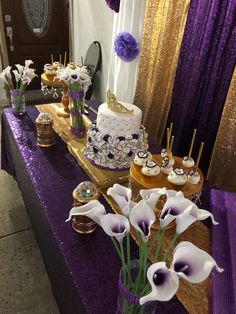 a table topped with purple and gold desserts covered in frosted cake next to flowers