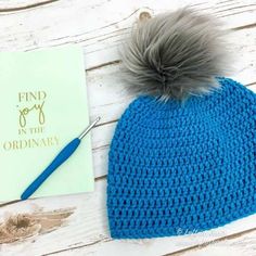 a blue crocheted beanie hat next to a book with a pom - pom on it