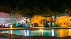 an outdoor pool at night with palm trees and people sitting around the poolside area