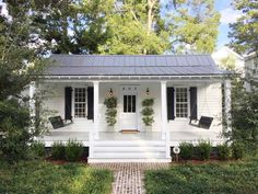 a white house with black shutters and two chairs on the front porch is surrounded by greenery