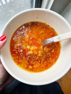 a person holding a bowl of soup with a spoon in it on a window sill