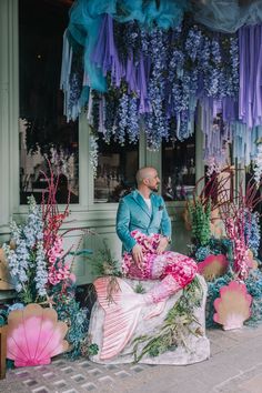 a man sitting in front of a flower shop with flowers hanging from the ceiling and decorations around him