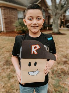 a young boy holding a cardboard box with a face drawn on it and wearing a hat