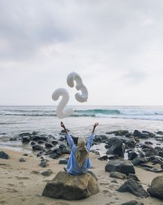 a woman sitting on top of a rock next to the ocean holding up two white numbers