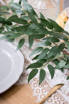 the table is set with white plates and greenery on top of lace doily