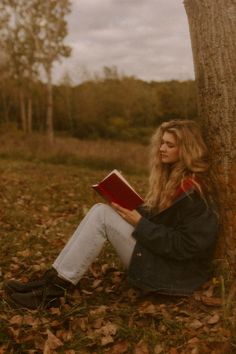 a woman is sitting under a tree reading a book in the fall leaves while looking at the camera