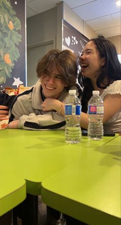 two people sitting at a green table with bottled water in front of them and smiling