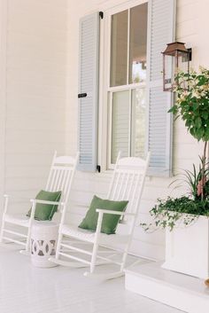 three white rocking chairs sitting on a porch next to a window with shutters and potted plants