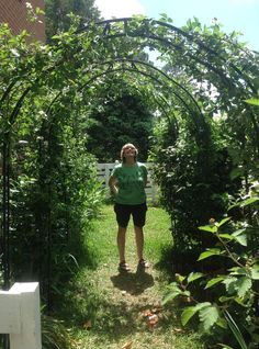 a woman standing in the middle of a garden with an arch over her head and bushes around her