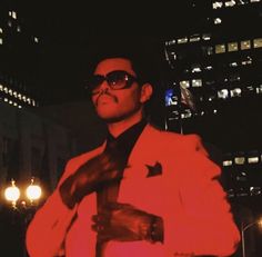 a man wearing sunglasses and a red jacket in the city at night with his hands on his chest