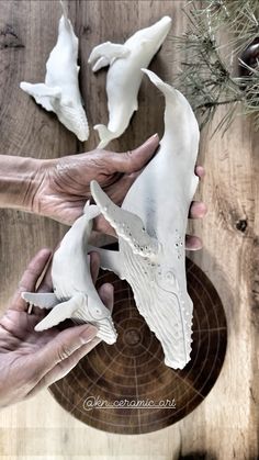 two hands are holding white sculptures on a wooden surface with pine needles and branches in the background