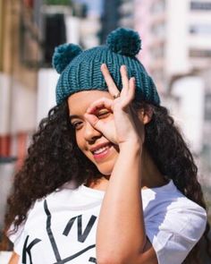 a woman with curly hair wearing a white shirt and a green knitted hat making the peace sign