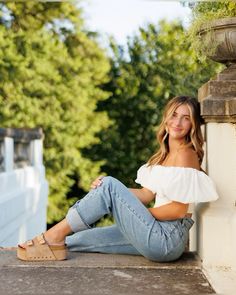 a beautiful young woman sitting on the side of a building wearing jeans and a white top