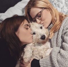 two women are kissing while holding a small white dog in their lap on the bed