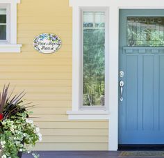 a blue front door on a yellow house with flowers in the foreground and a welcome sign above it